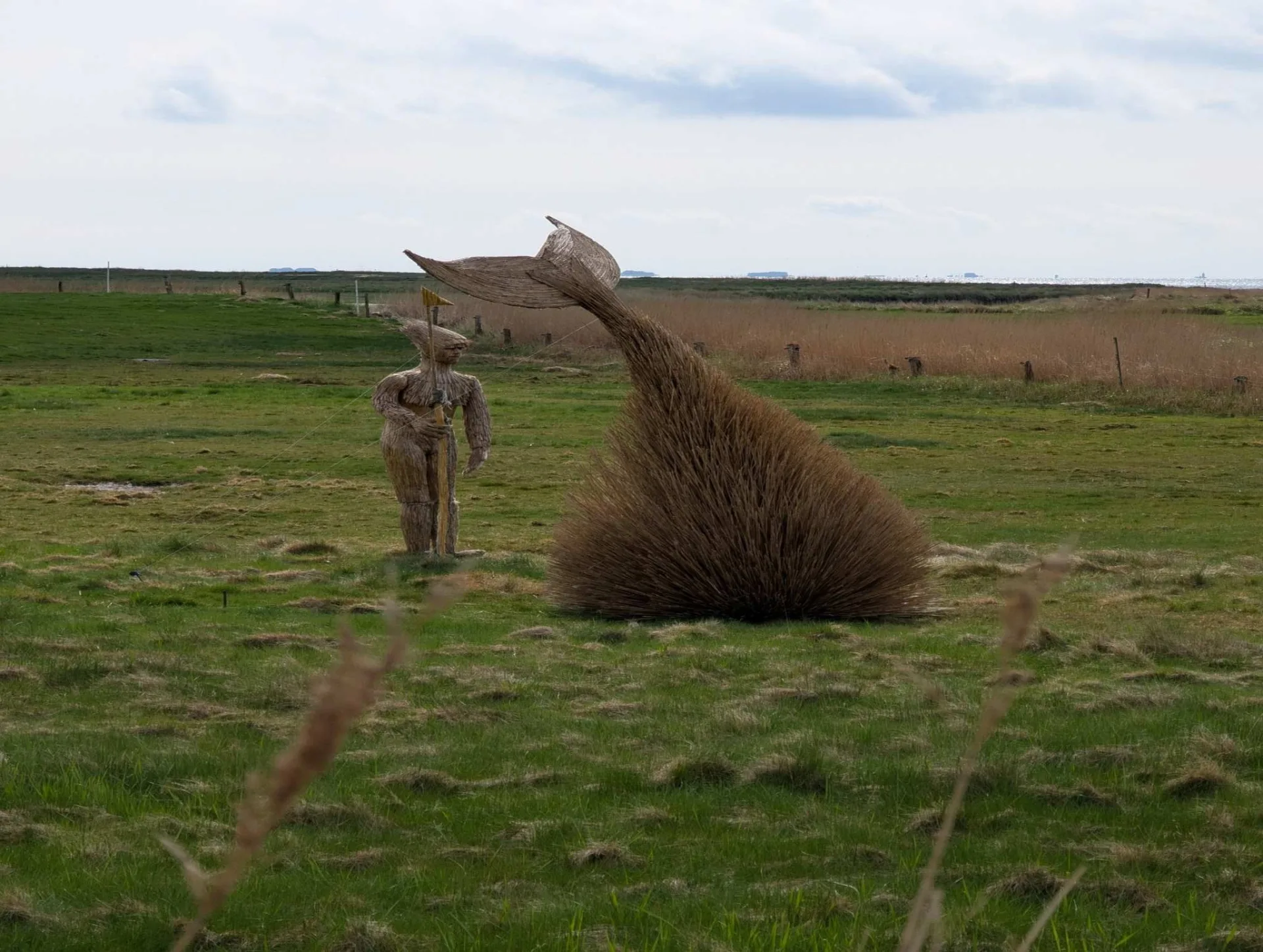 Landart in den Salzwiesen der Godel: ein abtauchender Wal aus geflochtenem Reet, daneben eine Figur – passend für Föhr, die alte Insel der Walfänger.